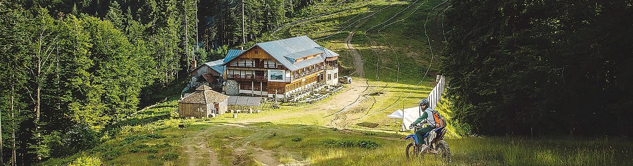 Toată proprietatea vara: cabana, foișorul, terasa cu mese și poteca prin iarbă.