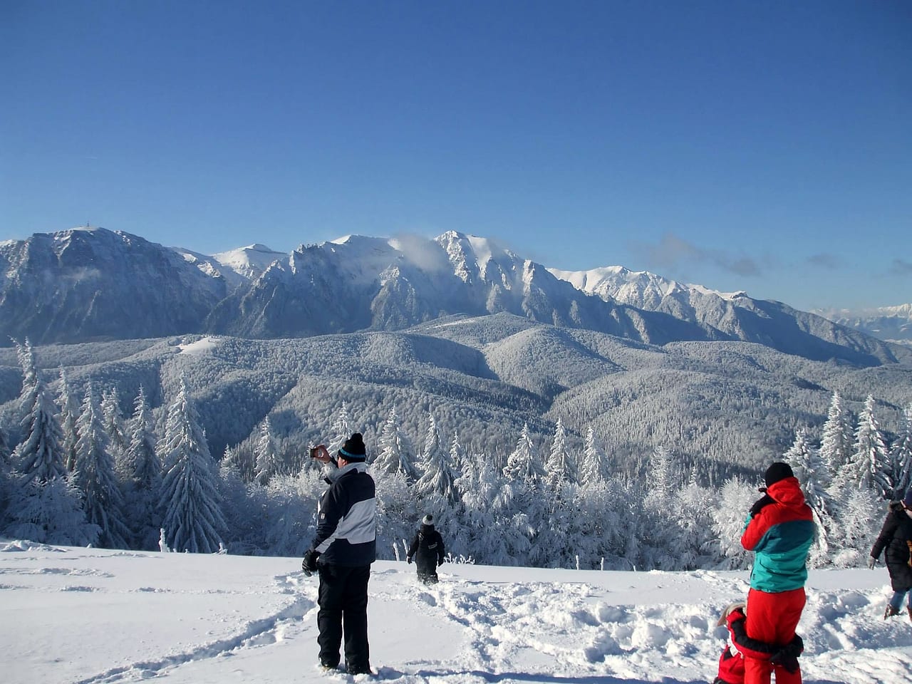 Panoramă spre Bucegi iarna, cu câțiva oameni care fotografiază de pe marginea poienii.