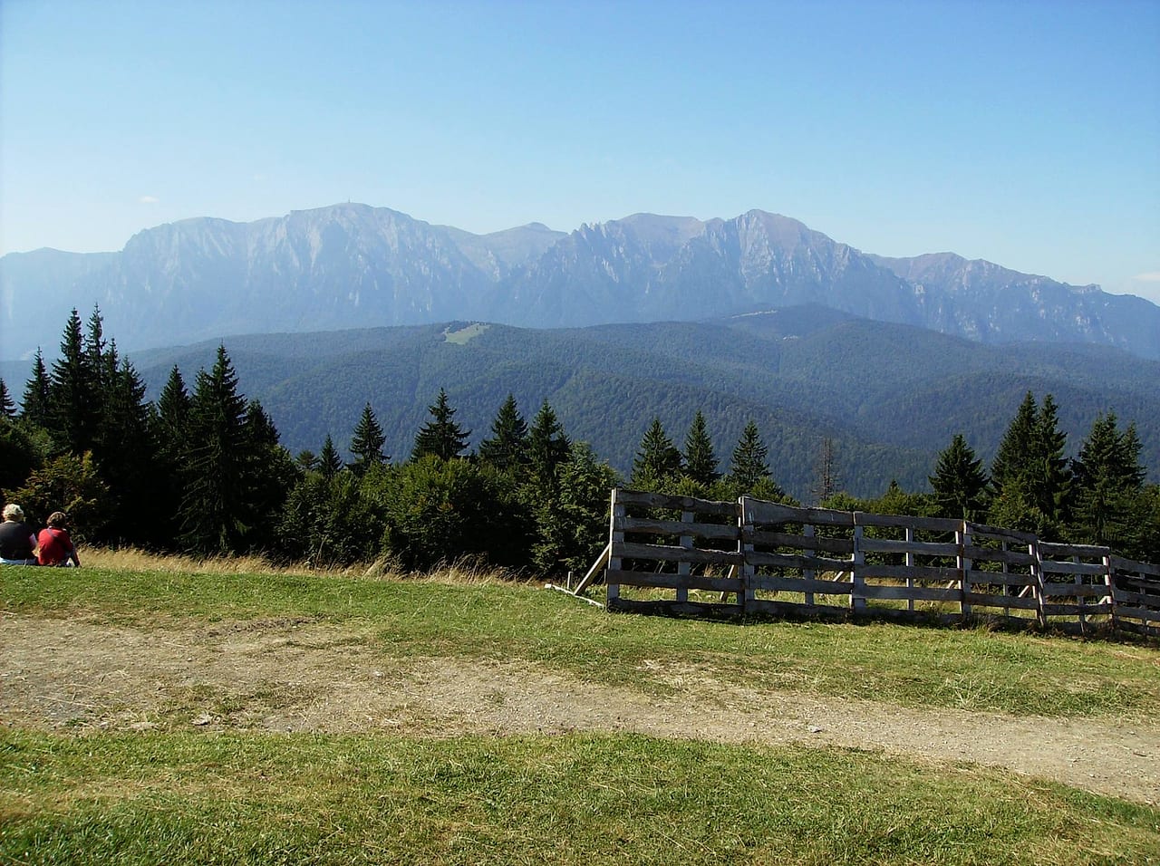 Priveliștea spre Bucegi de pe platou, cu un gard de lemn pe dreapta și doi drumeți așezați în iarbă.