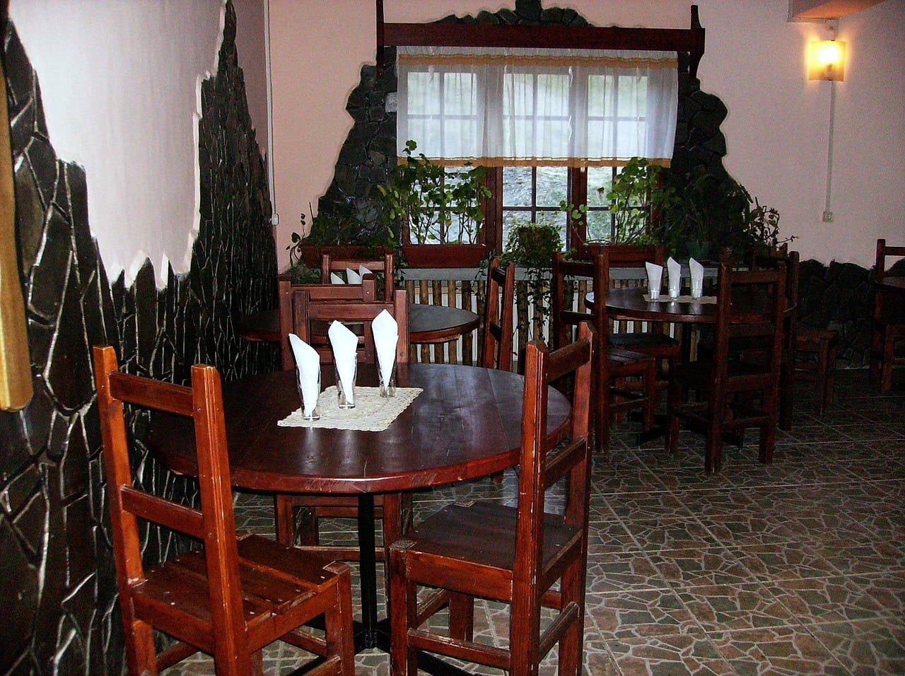 Sala restaurantului în lumină de zi, cu mese rotunde puse, perdele dantelate și peretele de piatră.
